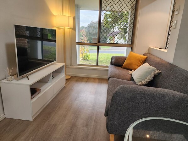 Ein modernes Wohnzimmer mit einem Flachbildfernseher auf einem weißen Standfuß, einem grauen Sofa mit Kissen und einem Fenster mit Blick ins Freie.