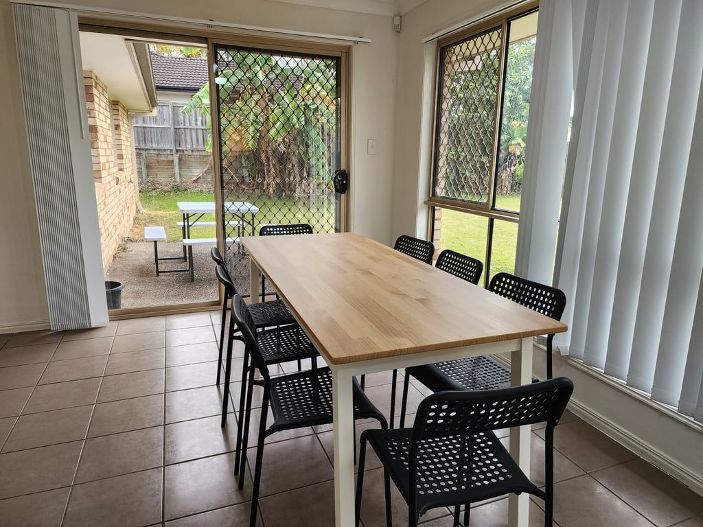 A dining area with a wooden table and black chairs, a sliding glass door leading to a backyard, and a view of a fence and greenery.