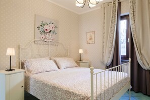 A bedroom with a bed, two bedside tables, a nightstand, a wall-mounted lamp, a window with curtains, and a floral painting. - Villa Sinfonia, Exclusive Villa in Ravello (Ravello)