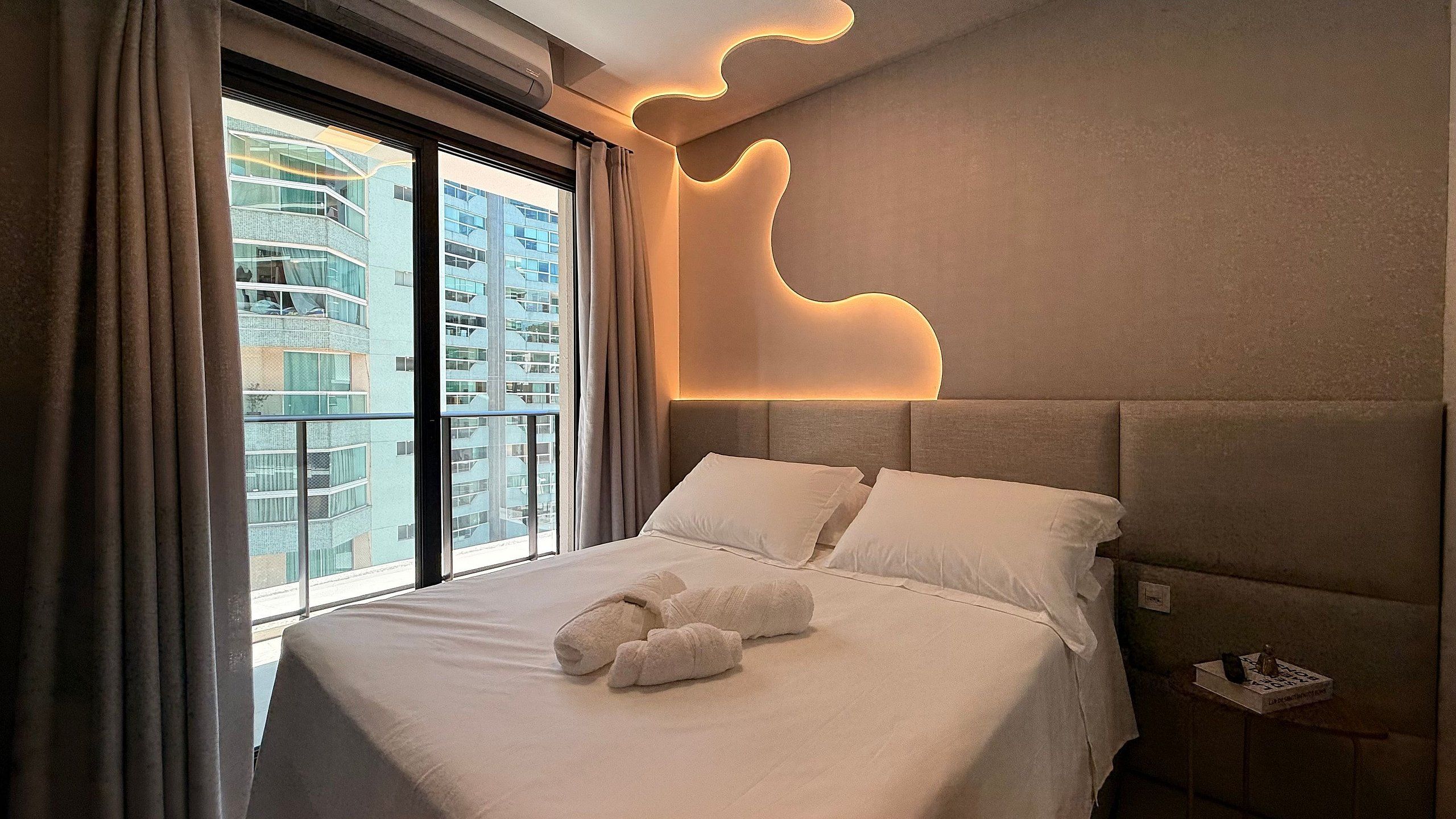 A hotel room with a large bed, a headboard with integrated lighting, and a view of a high-rise building.