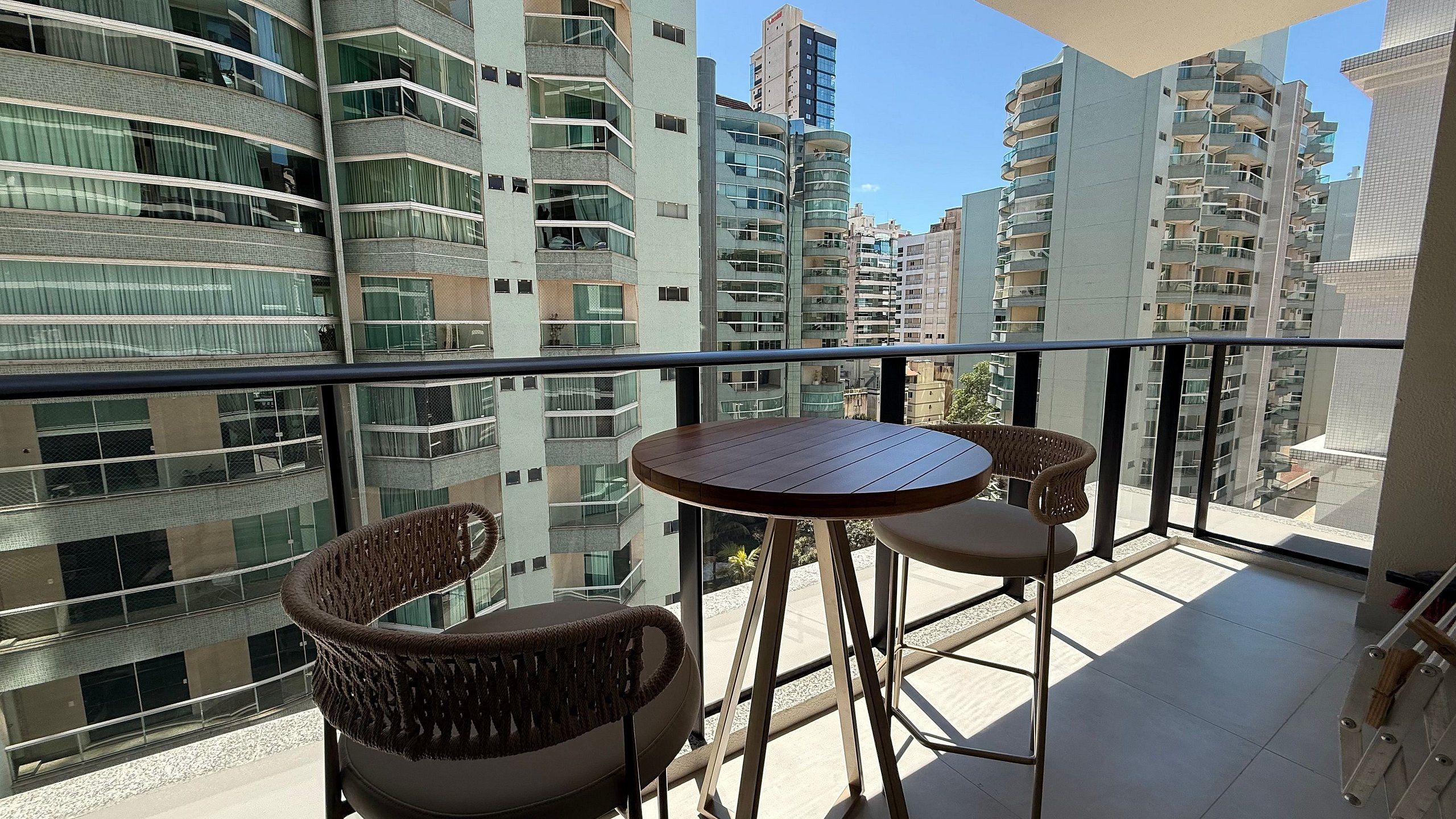 A balcony with a round table and two wicker chairs, overlooking a cityscape with high-rise buildings.
