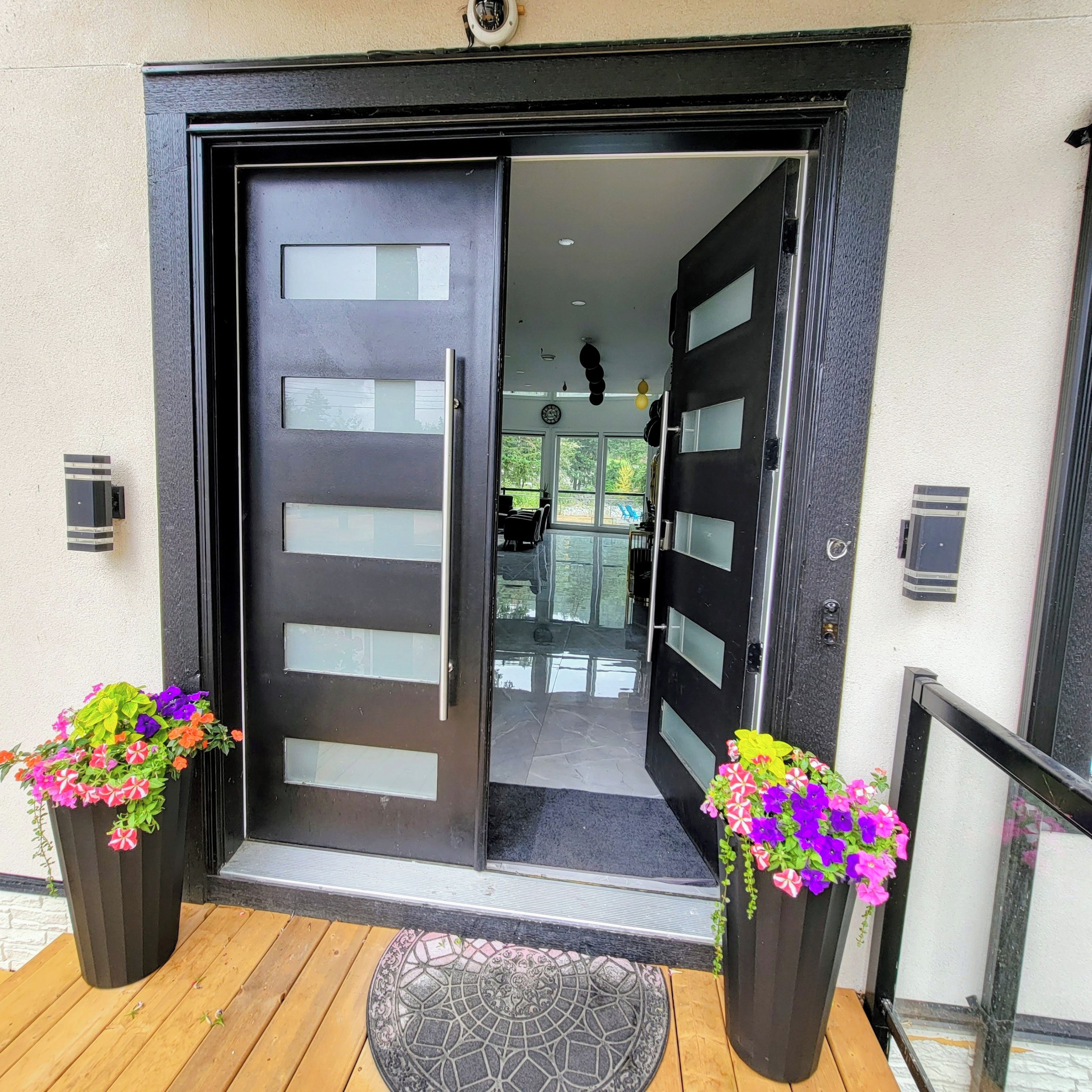 A modern entrance with a black door featuring horizontal panels and a glass panel. There are two flower planters on either side of the door.