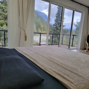 A bedroom with a large bed, a view of a mountainous landscape through floor-to-ceiling windows, and a balcony with a railing.