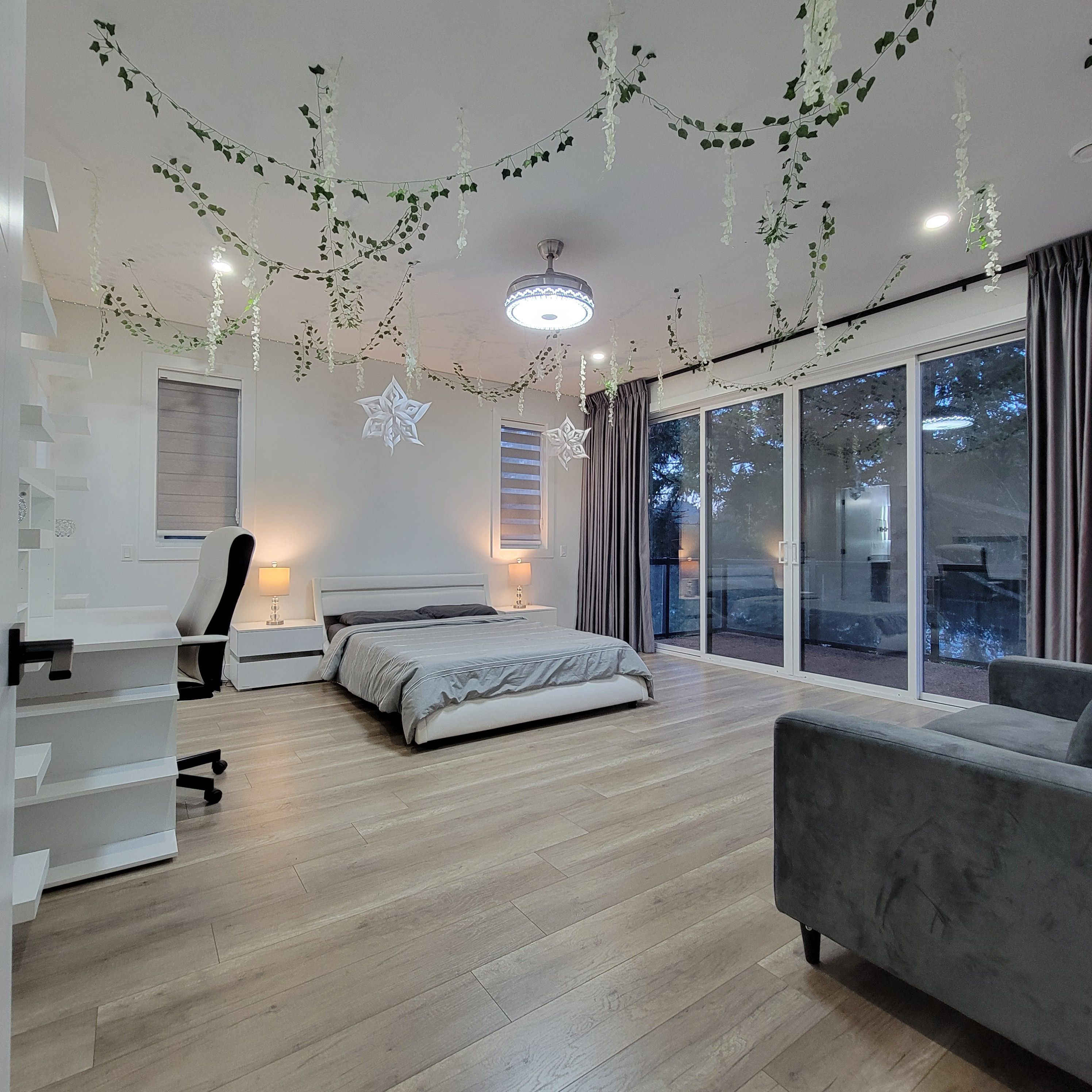A modern bedroom with a bed, a desk, and a chair. There is a large window with curtains and a view of the outside.