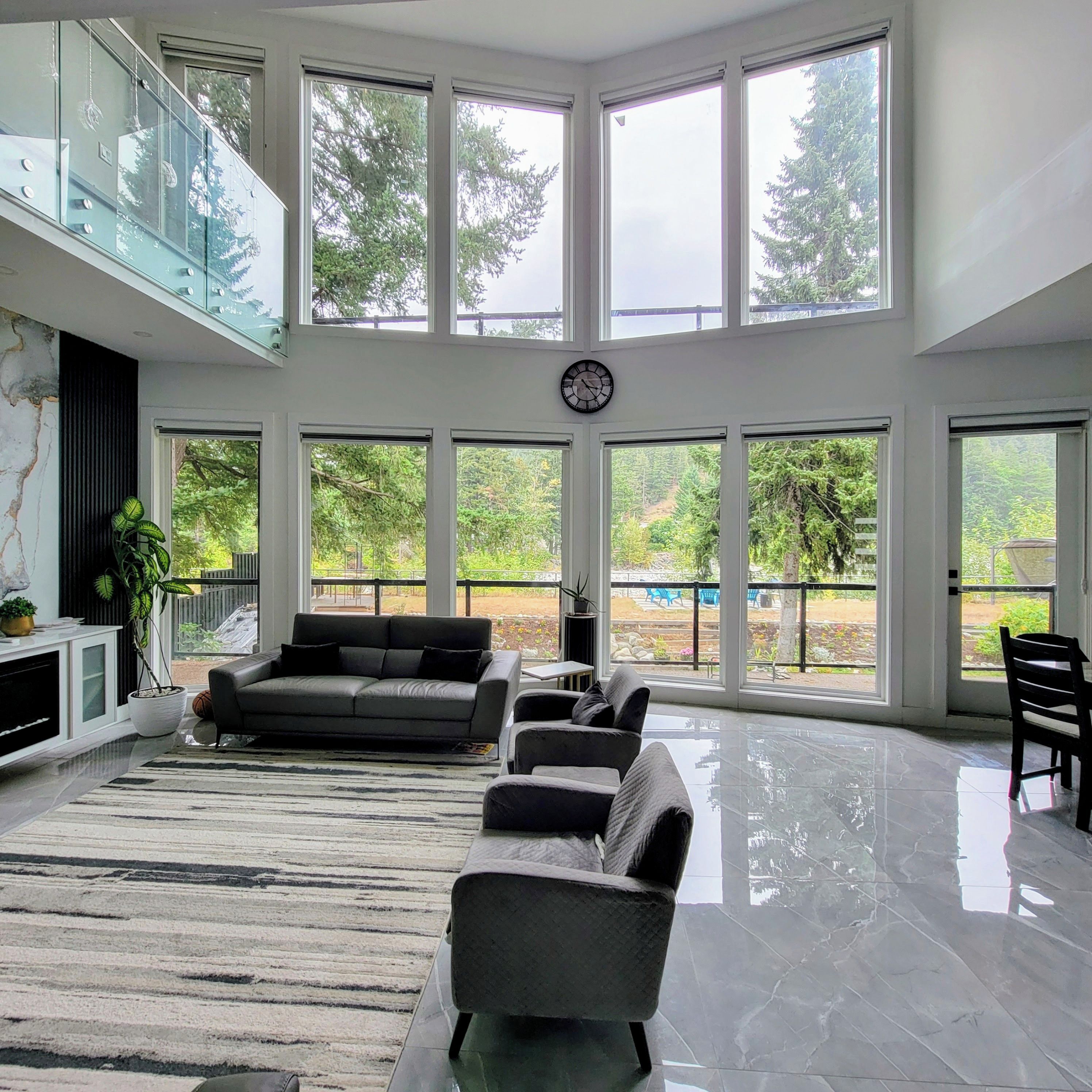 A spacious living area with a high ceiling, large windows, a sofa, a chair, and a clock on the wall.