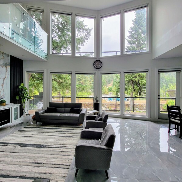 A spacious living area with a high ceiling, large windows, a sofa, a chair, and a clock on the wall.