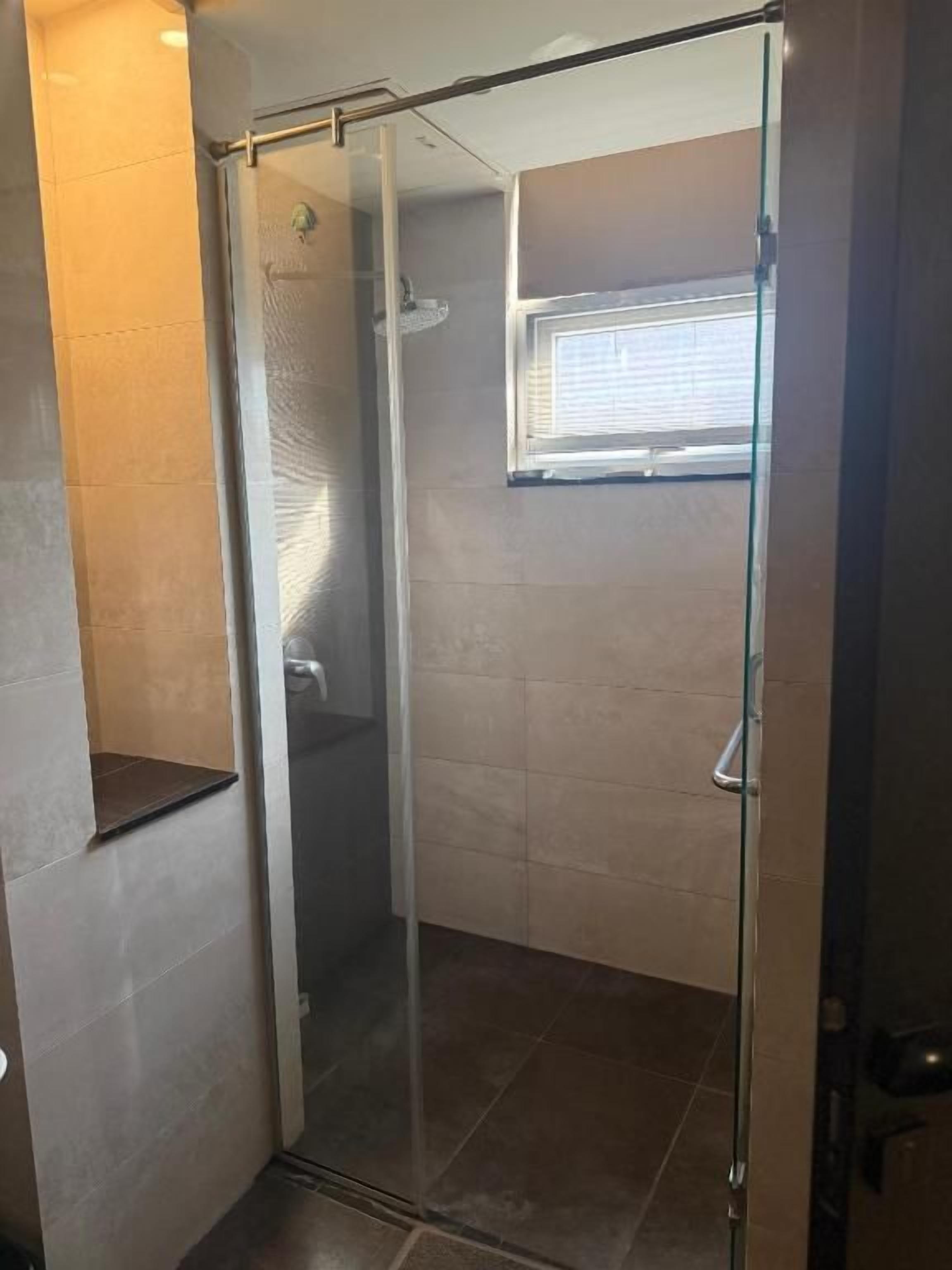 A glass-enclosed shower stall with a window above, tiled walls, and a door to the left.