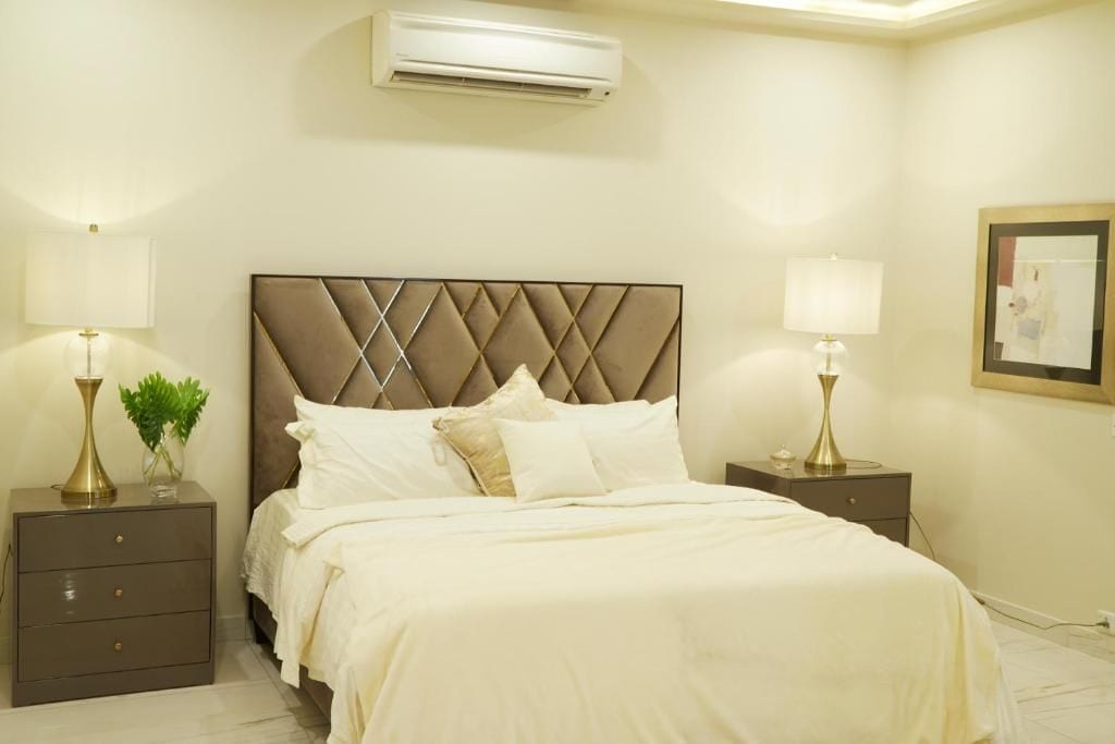 A hotel room with a bed, two bedside tables, lamps, a wall-mounted air conditioner, and a framed picture on the wall.