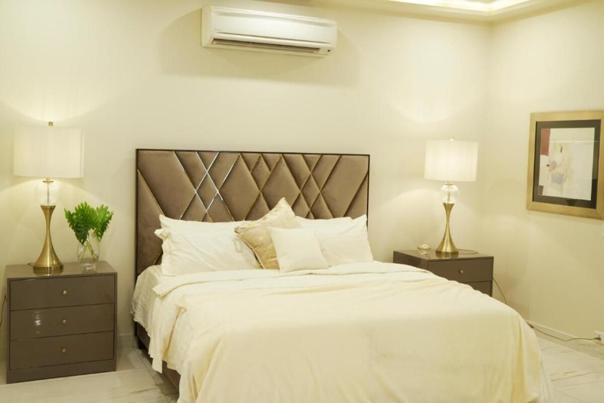 Habitación de hotel con cama, dos mesitas de noche, lámparas, aire acondicionado de pared y un cuadro enmarcado.