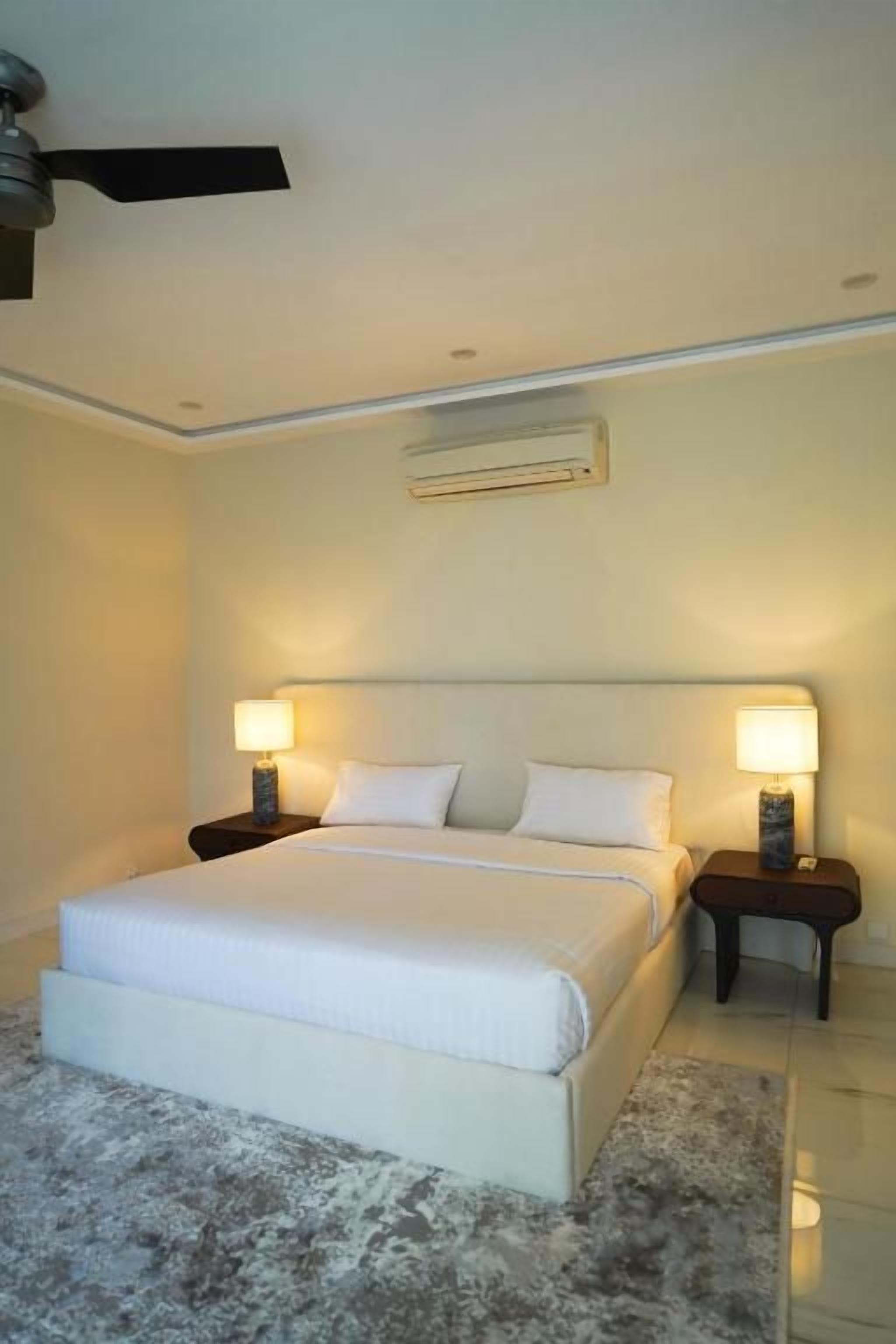 Un dormitorio con cama, dos mesitas de noche con lámparas, ventilador de techo y unidad de aire acondicionado.