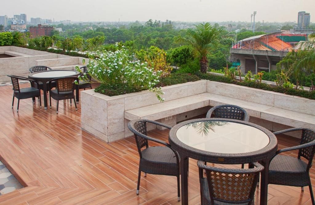 An outdoor seating area with round tables and chairs, surrounded by greenery and a cityscape view.