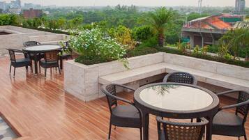 An outdoor seating area with round tables and chairs, surrounded by greenery and a cityscape view.