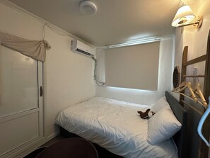 A small, single-bed room with a window, an air conditioning unit, a bedside table, and a hanging light fixture.