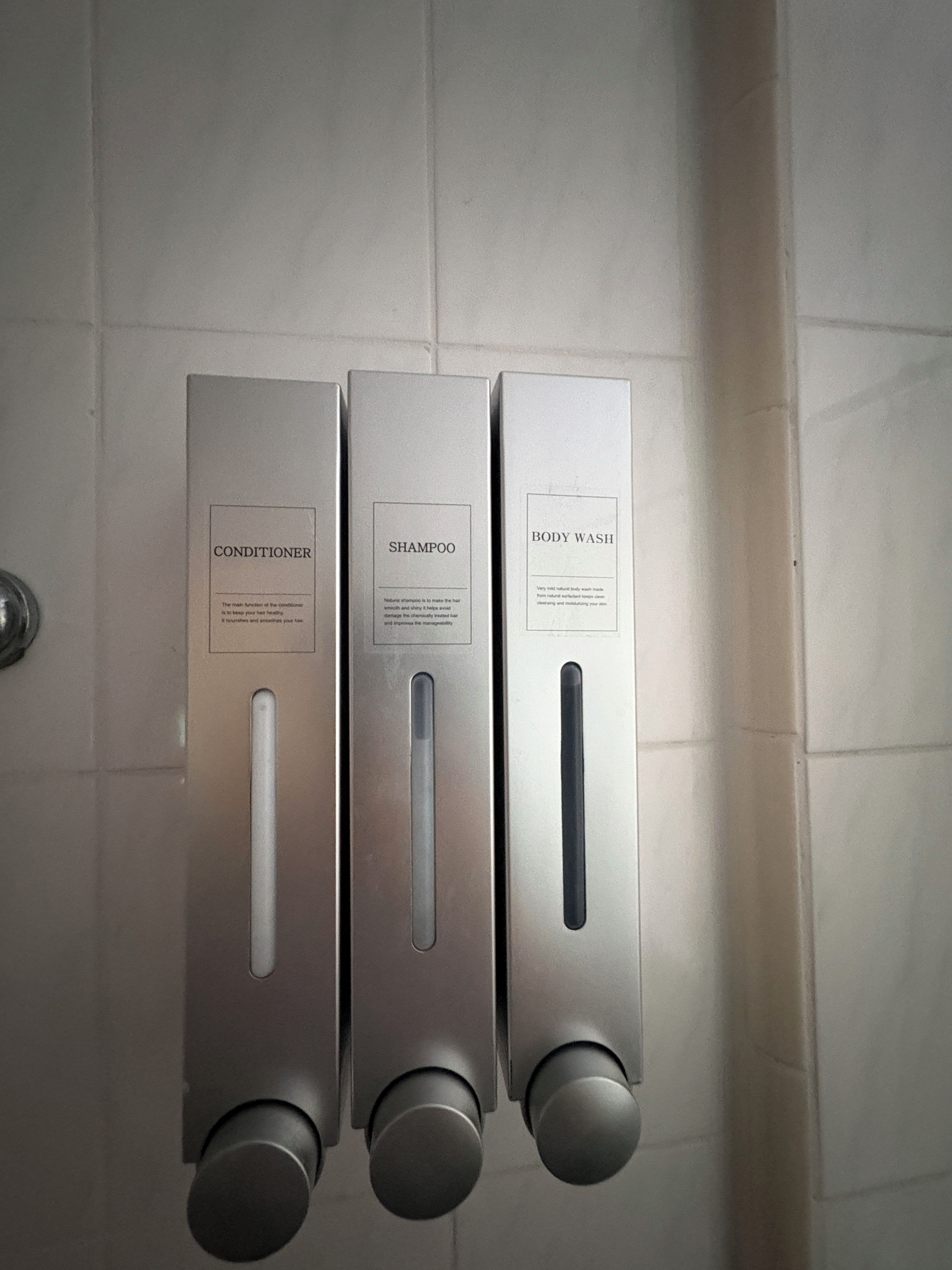 Three metallic dispensers labeled conditioner, shampoo, and body wash mounted on a tiled wall.