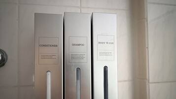 Three metallic dispensers labeled conditioner, shampoo, and body wash mounted on a tiled wall.