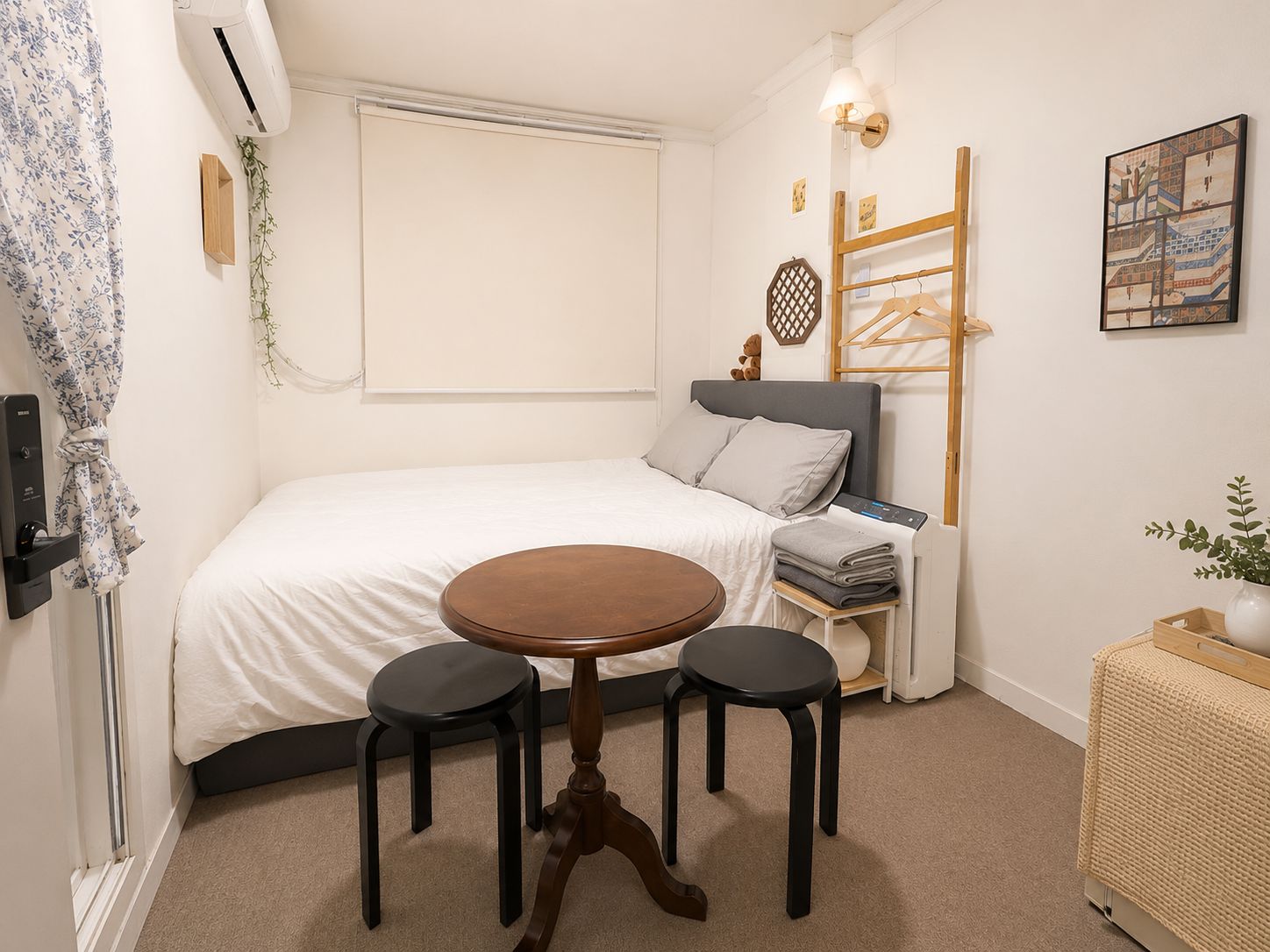 A compact bedroom with a bed, a round wooden table with two stools, a bedside table with a lamp, and a wall-mounted shelf with a teddy bear.