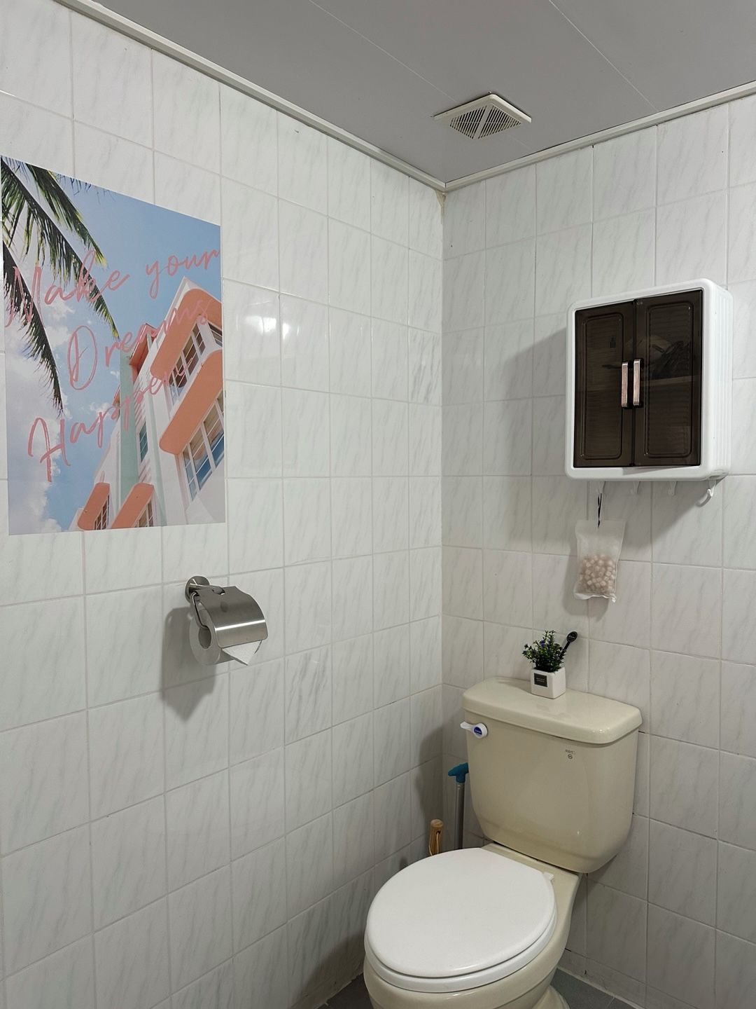 A bathroom with white tiles, a toilet, a hand dryer, a cabinet, and a poster on the wall.