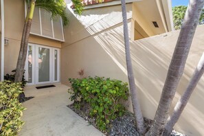 A residential building with a sliding glass door entrance, a concrete pathway, and a small garden area with plants and a palm tree. - Fantastic 2-bedroom condo in soothing Palm Beach Gardens (Palm Beach Gardens)