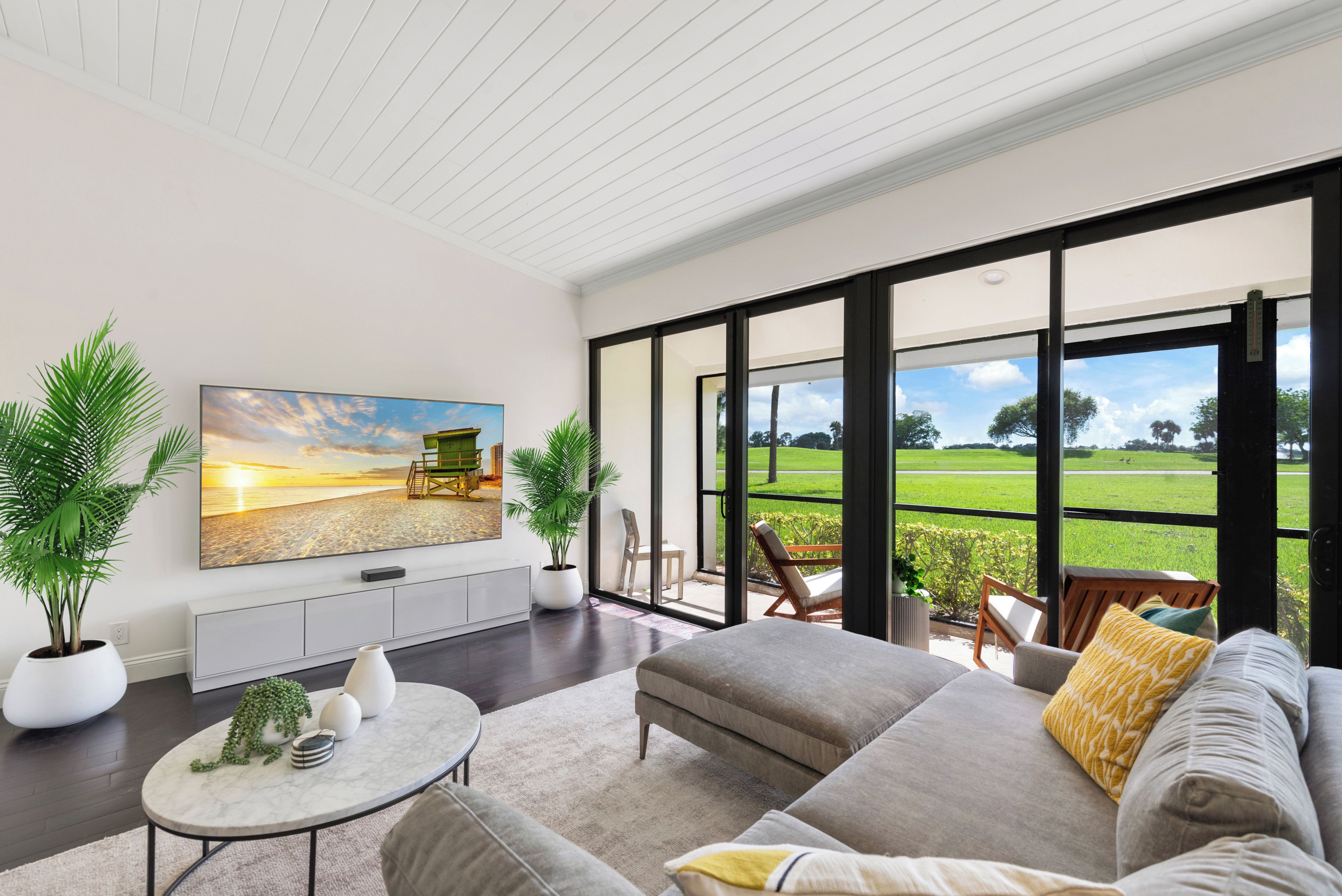 A modern living room with a large TV, comfortable seating, and a view of a green landscape through large windows.
