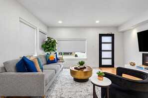 Un salon moderne avec un canapé gris, une table basse ronde et un fauteuil noir. Une grande fenêtre donne sur la rue, une porte à cadre noir et un téléviseur à écran plat est fixé au mur.