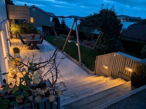 A wooden deck with a table and chairs, surrounded by greenery and a view of a body of water.