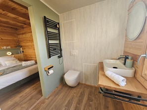 A compact bedroom with a bed, a bedside table, a wall-mounted heater, a toilet, and a sink with a mirror.