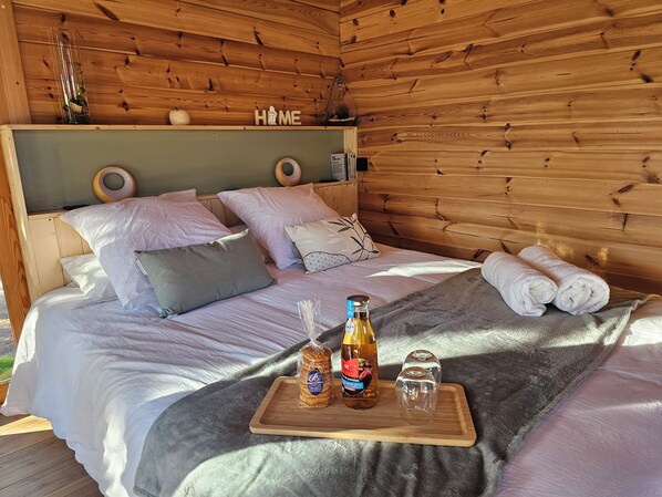 A wooden cabin bedroom with a bed, pillows, a tray with bottles, and a 'HOME' sign on the wall.
