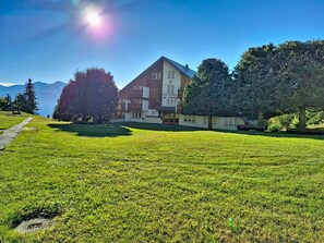 A hotel with a clear view of the mountains in the background, a well-maintained lawn, and a pathway leading to the entrance. - Le Chamois d'Or A303 by Interhome (Crans-Montana)