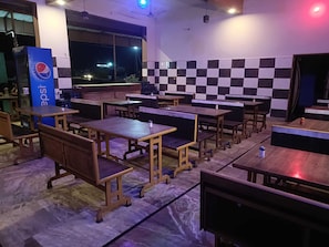 A restaurant interior with multiple wooden tables and benches, a Pepsi vending machine, and a checkered wall.