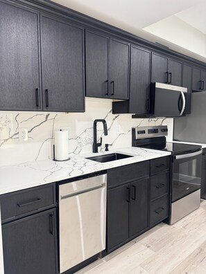 A modern kitchen with dark cabinets, a white marble countertop, a stainless steel dishwasher, and a microwave. - Modern 2 Bedroom Studio Retreat Near Maple-Lawn (Fulton)