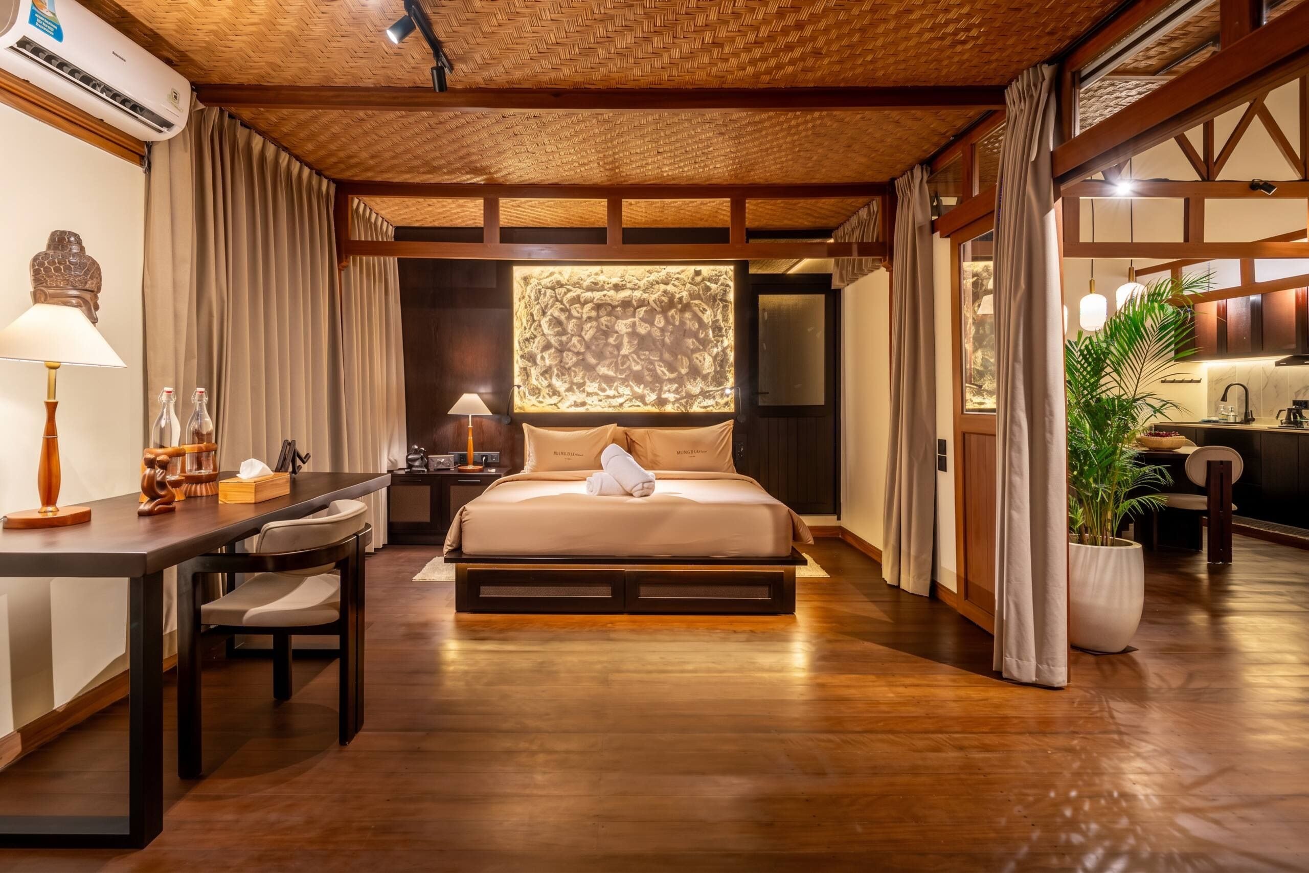 A hotel room with a bed, desk, and chair, a potted plant, and a view of the outside.