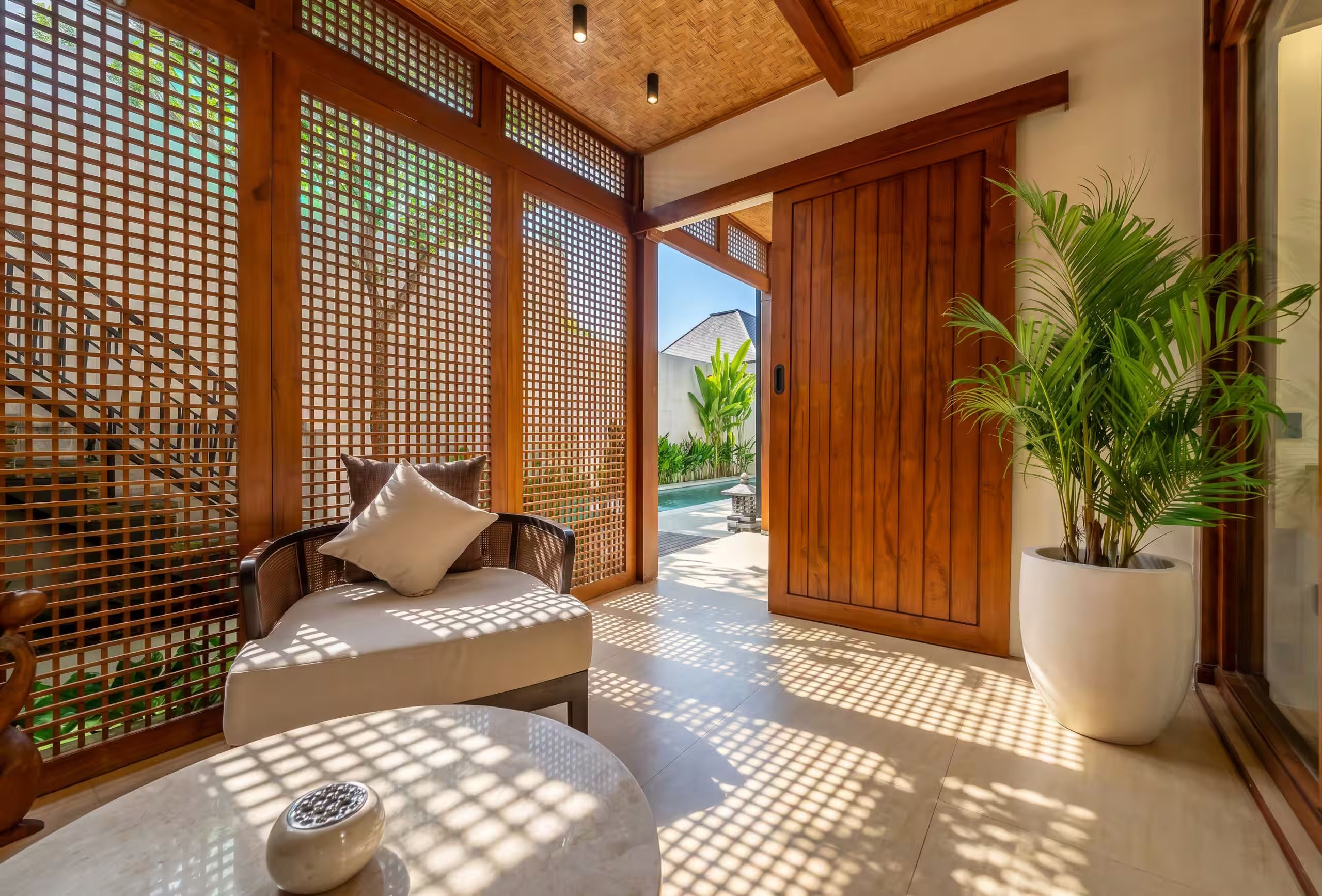 Una habitación con un separador de madera, una zona de estar con cojines, una planta en maceta y vistas a una piscina.