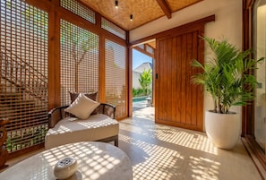 Una habitación con un separador de madera, una zona de estar con cojines, una planta en maceta y vistas a una piscina.