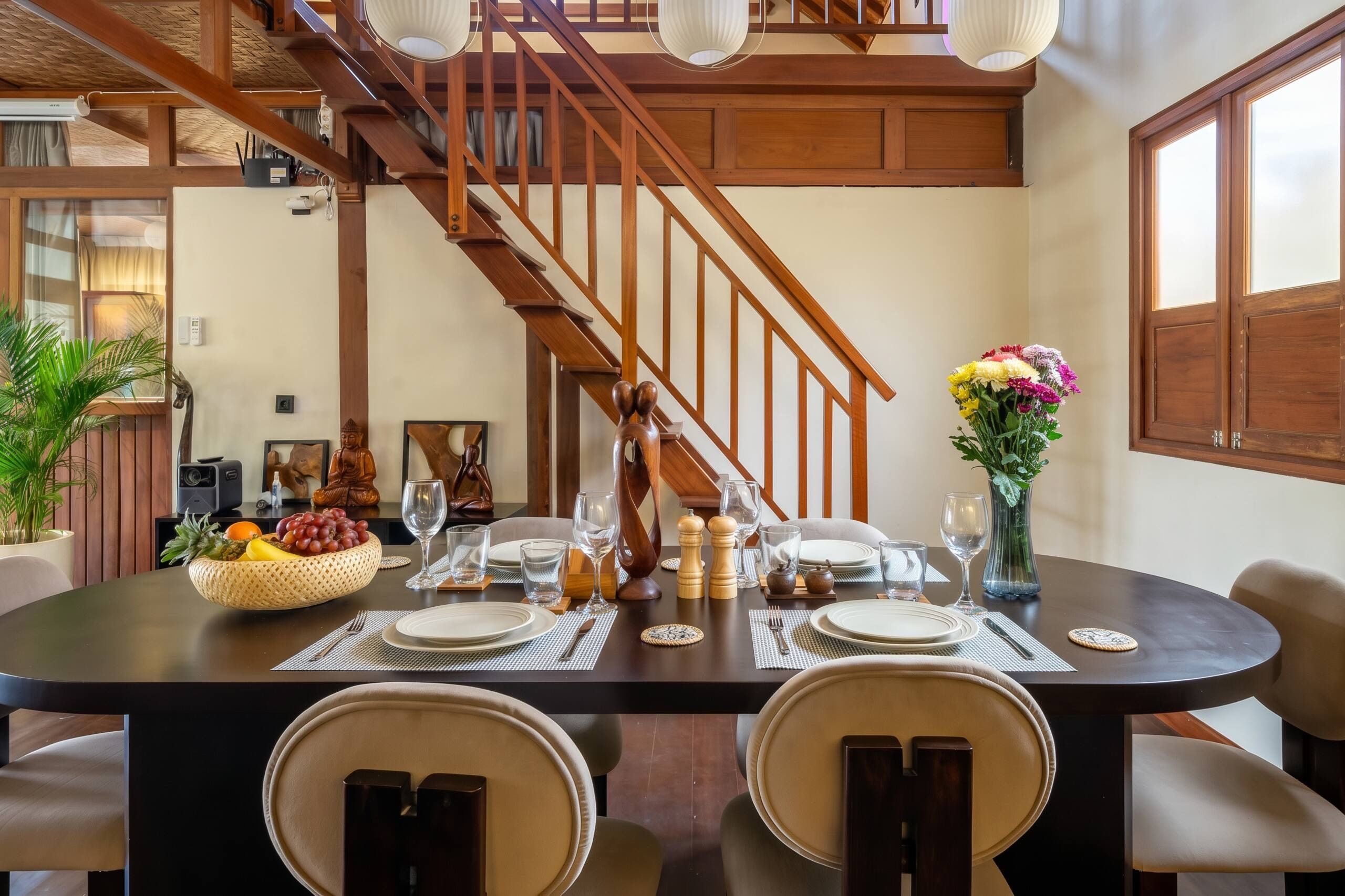 Un comedor con una escalera de madera, un jarrón de flores y un frutero sobre la mesa.