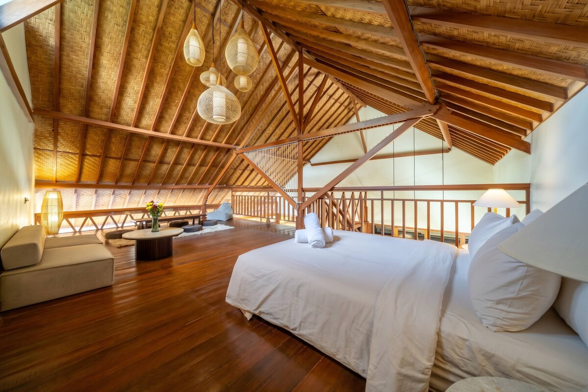 Een ruime kamer met een hoge nok, houten balken, een bed met witte lakens, een bank en een bijzettafeltje.