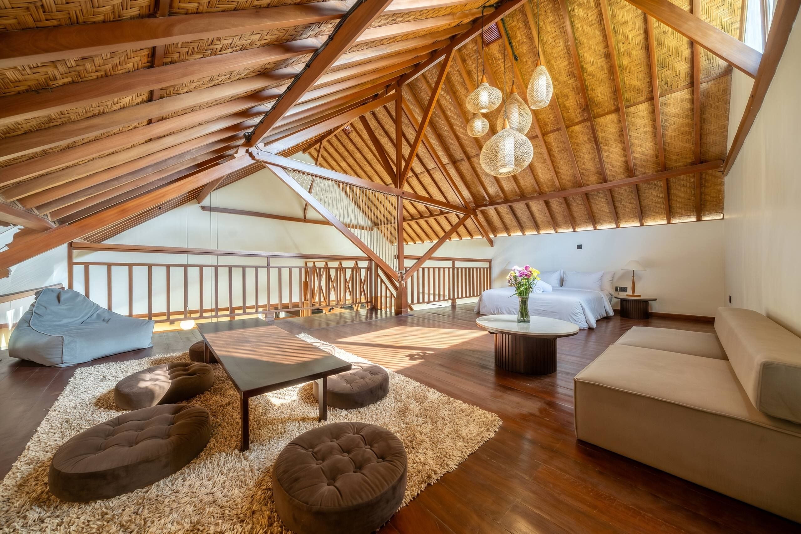 A spacious loft with a high ceiling, wooden beams, a mezzanine level, and a cozy seating area with a sofa, ottoman, and bean bag.