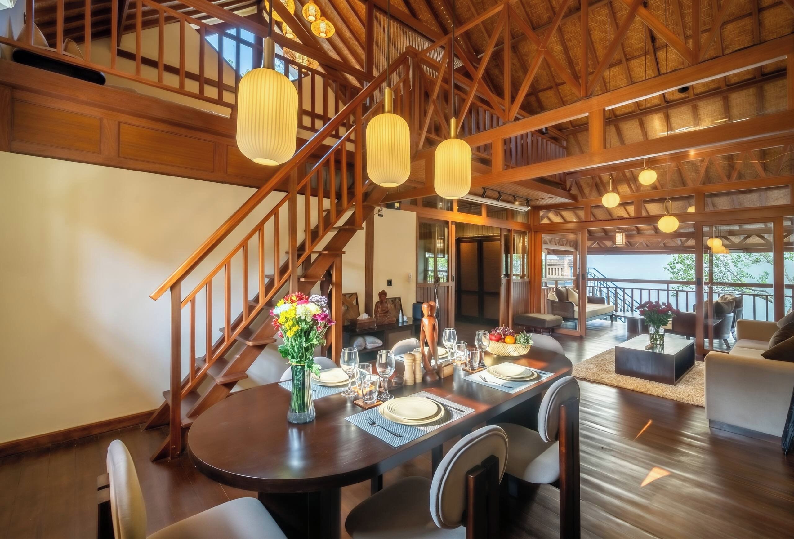 A dining area with a wooden staircase, a dining table set for four, and a view of the outdoors through large windows.