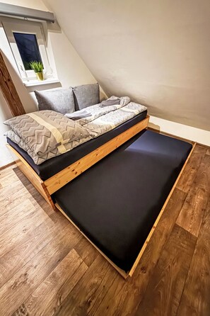 Uma cama estilo loft com estrutura de madeira e colchão preto, em um quarto com piso de madeira e uma pequena janela com uma planta.