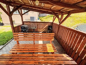 Een houten bank onder een pergola, met een barbecue en een tafel.