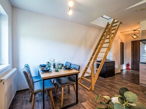 Ein Essbereich mit Holztisch, gedeckt für zwei Personen, einer an der Wand stehenden Leiter und Blick in die Küche im Hintergrund.
