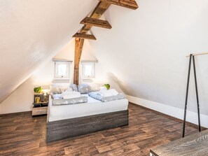 Ein Schlafzimmer mit schrägverdeckter Decke, einem Bett mit maritimer Bettwäsche, einem Nachttisch mit Lampe und Blick durch ein Fenster.