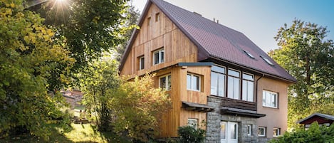 A two-story wooden house with a sloped roof, surrounded by lush greenery and a stone pathway leading to the entrance.