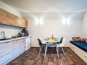 Een kleine keuken met houten kastjes, een magnetron en een spoelbak. Er staat een eettafel met stoelen en een kleine bank met kleurrijke kussens.