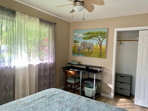 A bedroom with a bed, a desk, a chair, a ceiling fan, a painting of elephants, and a closet. - Beautiful Hale Close to the Ocean (Pāhoa)