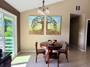 Uma área de refeições com uma mesa redonda, duas cadeiras e uma tigela de frutas. Há duas pinturas penduradas na parede, retratando árvores com rostos humanos.