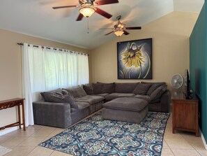 A living room with a sectional sofa, a ceiling fan, a wall-mounted painting, and a patterned rug. - Beautiful Hale Close to the Ocean (Pāhoa)