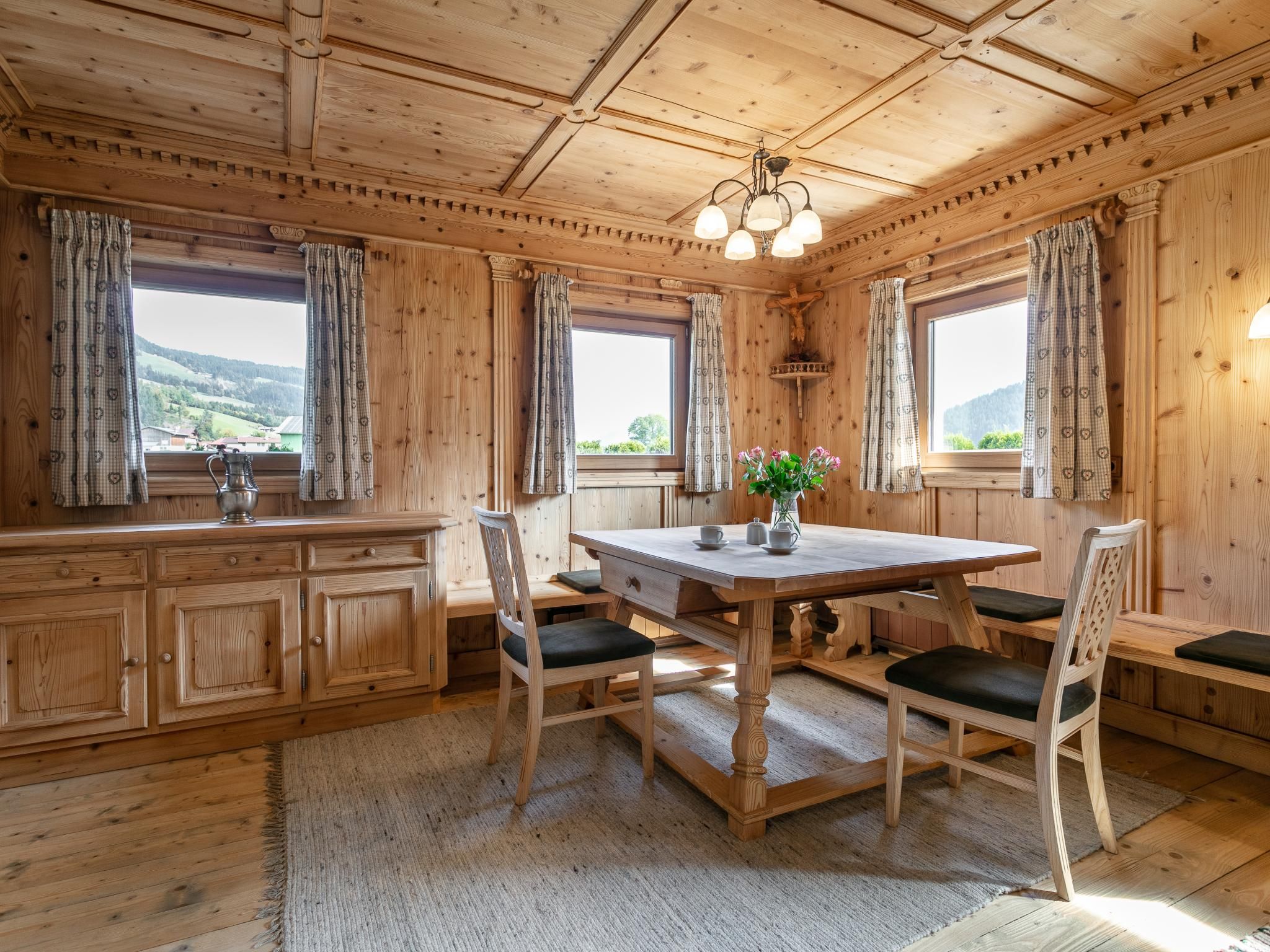 A wooden interior with a dining table, chairs, a chandelier, and windows with curtains.