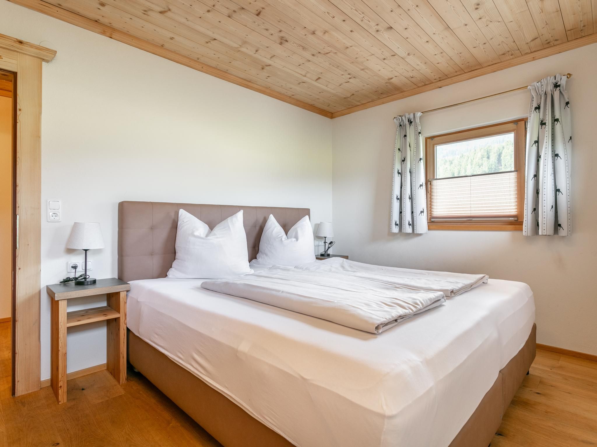 Una camera da letto con un letto, un comodino, una lampada, una finestra con tende e un soffitto in legno.