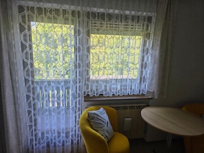 Quarto com duas janelas cobertas por cortinas de renda branca, uma cadeira amarela e uma pequena mesa redonda.