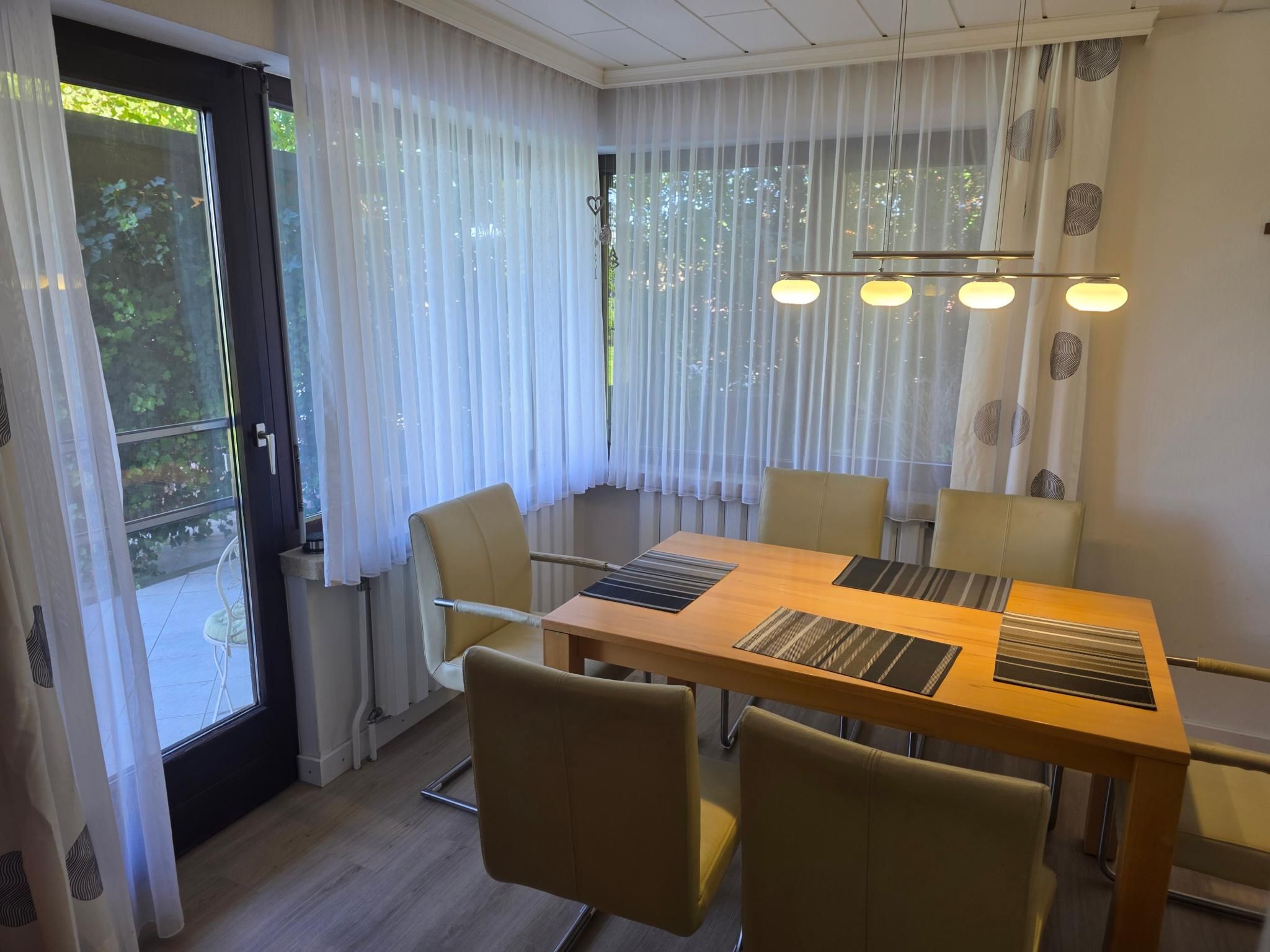 A dining area with a wooden table and chairs, a window with sheer curtains, and a ceiling light fixture.